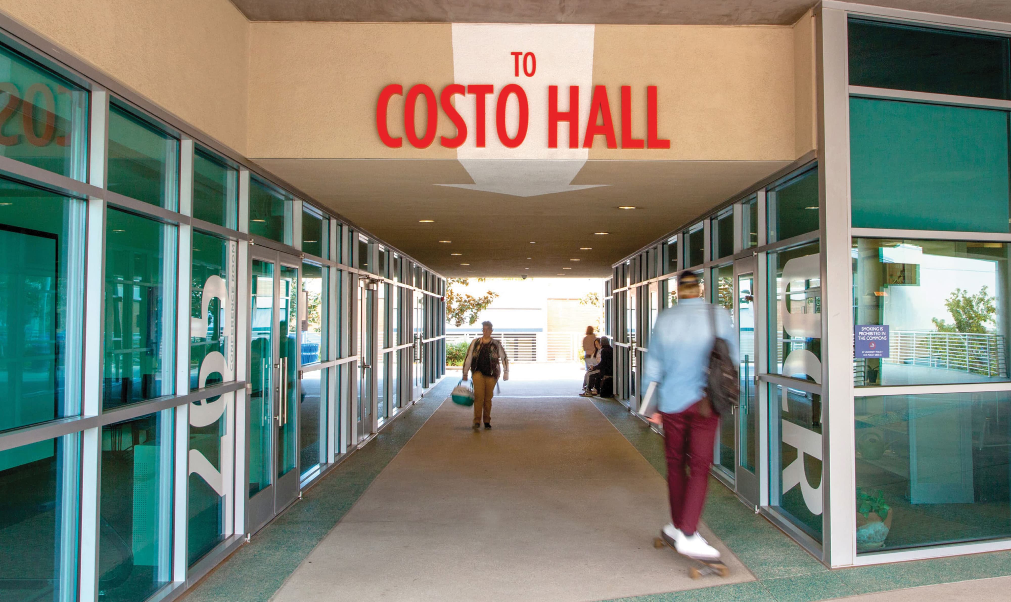 Covered walkway between glass-walled buildings with a red 'To Costo Hall' directional sign overhead, as a person skateboards through the passage and another walks toward the camera.