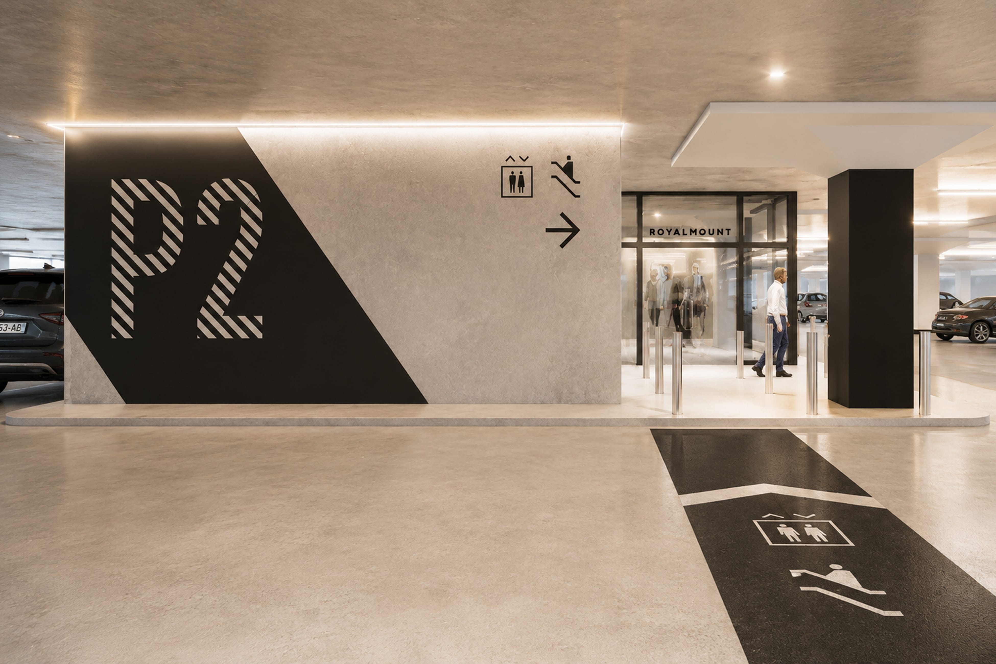 Parking garage entrance with large striped "P2" wall graphic, wall and floor pictogram signage for elevator and escalator, and glass Royalmount entry doors — parking wayfinding design by RSM Design