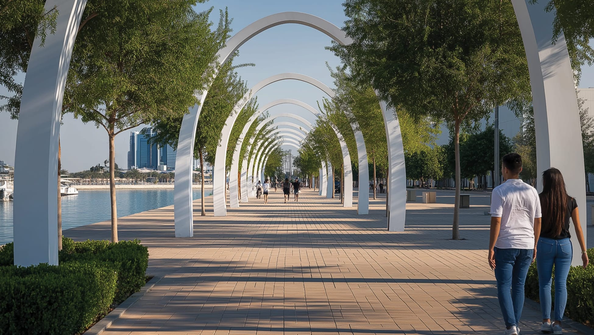 Ramhan Island Marina is a waterfront development in Abu Dhabi, UAE. The promenade shown features a signature series of white parabolic arches that frame a pedestrian walkway running parallel to the marina. The design integrates shade trees, low hedges, and warm-toned paving to create a comfortable, human-scale public realm suited to the Gulf climate. The skyline of Abu Dhabi is visible across the water, grounding the project within its urban context. It's a strong example of RSM Design's approach to landscape architecture that balances bold structural gestures with livable, activated public space.