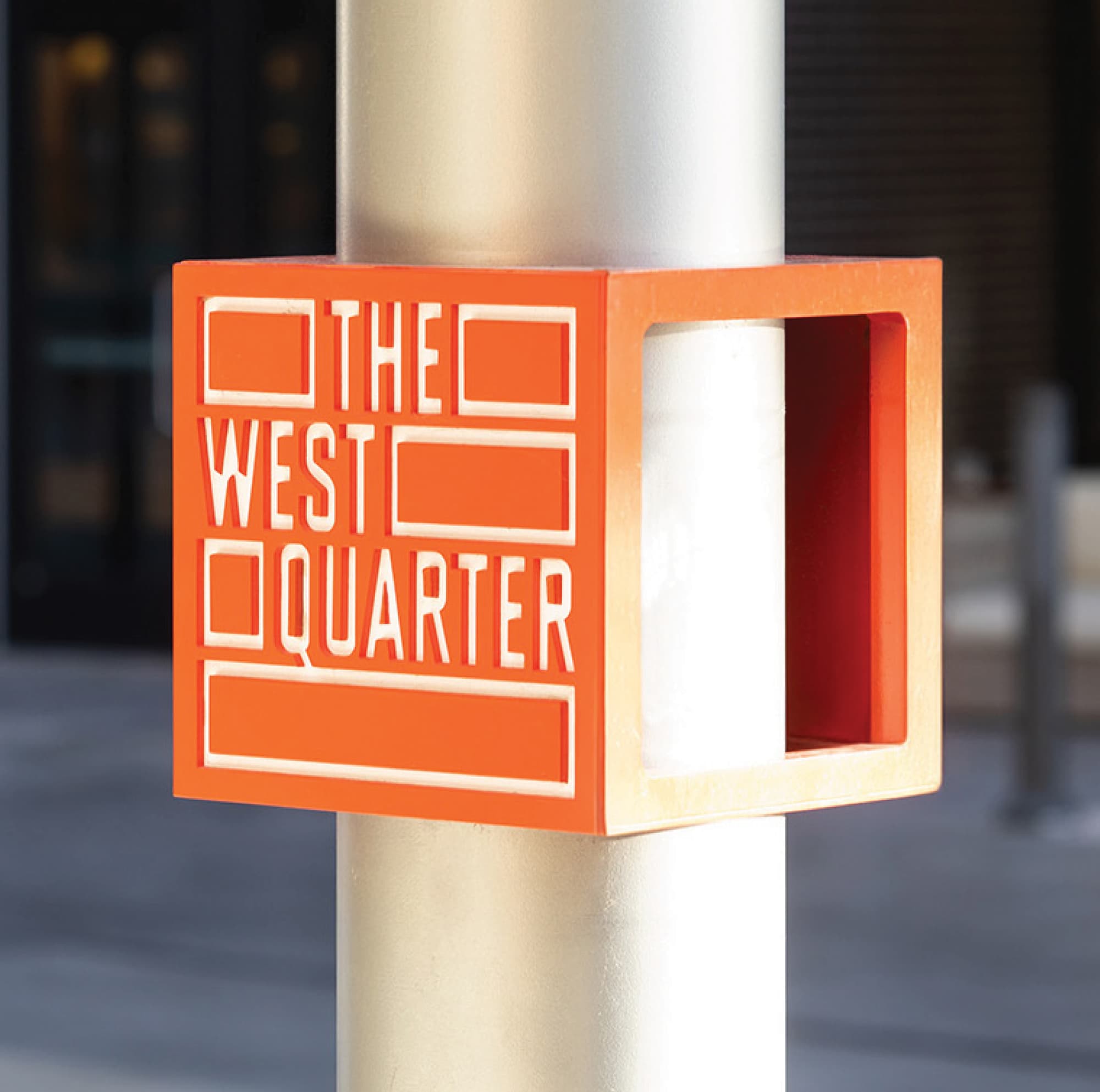 RSM Design street furniture branding and master sign program detail for The West Quarter in Salt Lake City, featuring a close-up of a three-dimensional orange cube pole collar with routed logo lettering and geometric brand pattern elements wrapping a gold streetscape light pole, revealing the orange interior on the open face, showcasing how environmental graphic design, specialty graphics, and visual identity extend into every layer of the pedestrian environment to reinforce a cohesive branded experience across mixed-use, residential community, hospitality, and streetscape markets.