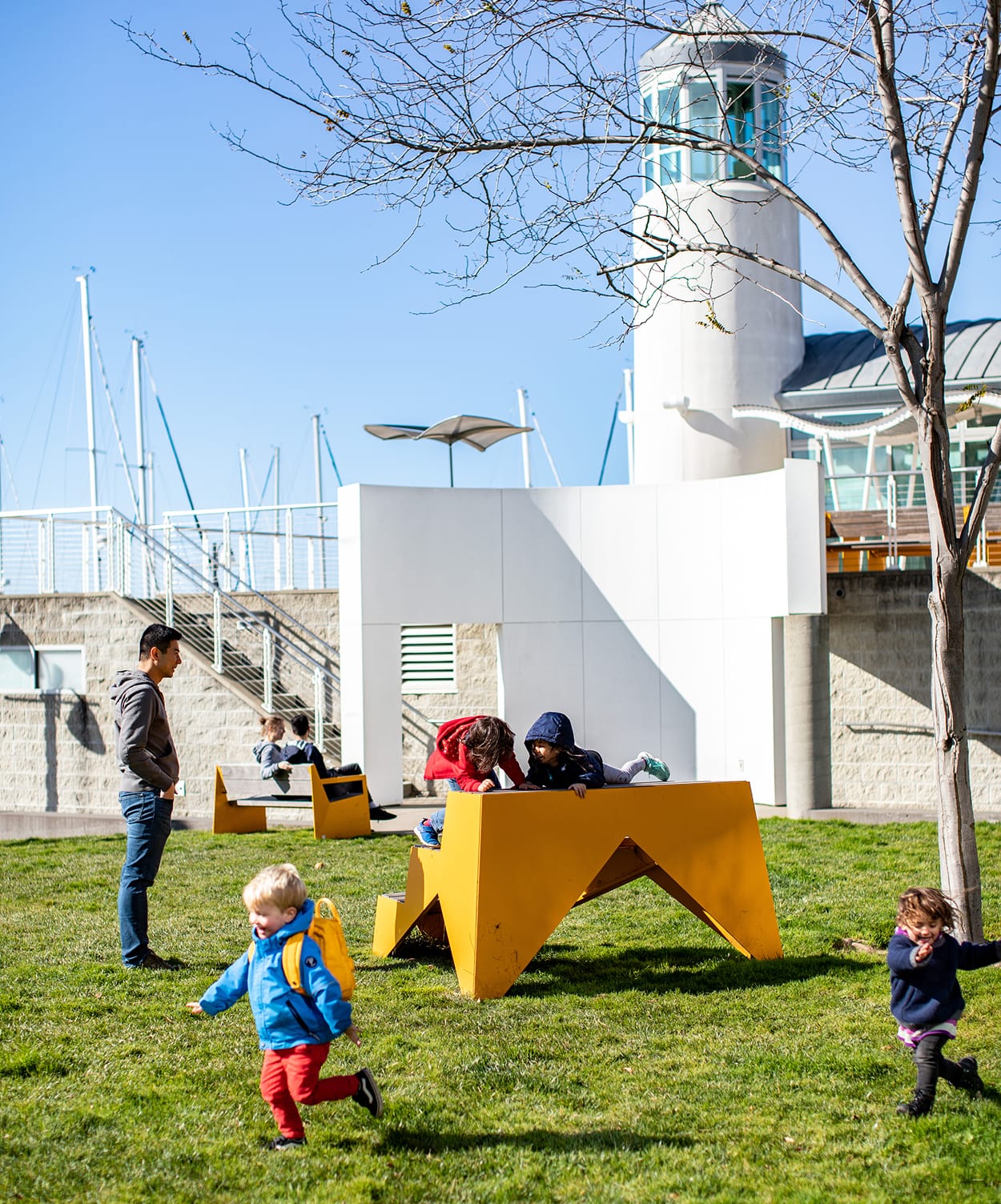 Families and children playing on yellow geometric outdoor furniture and green lawn at Jack London Square waterfront park with marina sailboats and lighthouse tower in background, Oakland, California